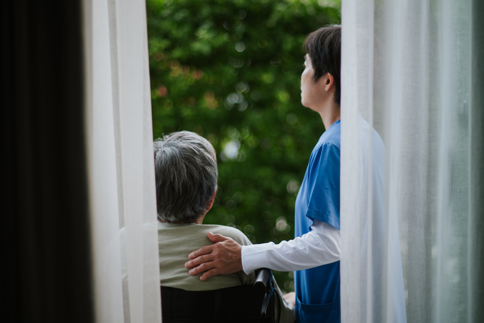 Elderly woman in wheelchair comforted by caregiver through curtain.